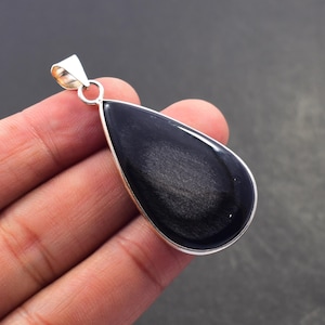Peut inclure: Un pendentif en forme de goutte d'eau avec une bélière argentée. Le pendentif présente une pierre noire polie avec des stries argentées. La pierre est sertie dans une lunette argentée. Le pendentif est tenu dans une main.