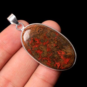 May include: Oval pendant with a silver-coloured bail. The pendant features a polished stone with a mix of red, brown, and green patterns. The stone is set in a silver-coloured bezel. The background is black.