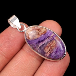 May include: Oval pendant with a polished charoite gemstone, displaying purple, brown, and white swirls. The stone is set in a silver bezel with a bail for a necklace. The pendant is held in a hand against a black backdrop.