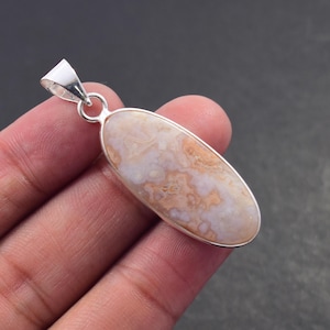 Puede incluir: Colgante ovalado con una piedra de color crema y melocotón engastada en un marco de metal plateado. La piedra tiene un aspecto veteado. El colgante tiene un lazo en la parte superior para una cadena.