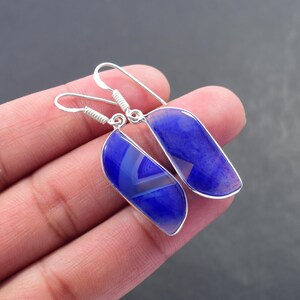 May include: A pair of blue agate earrings with a silver-coloured metal frame and ear wires. The earrings have a teardrop shape with a polished finish. The agate stone features a mix of light and dark blue hues with a banded pattern.