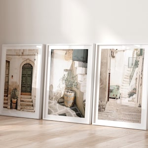 Op de afbeelding: Drie ingelijste kunstprints met Europese straatscènes. De prints tonen deuropeningen, trappen en een kleine auto in een smalle steeg. Het kleurenpalet is neutraal met beige, witte en groene tinten, wat een serene en klassieke esthetiek creëert.