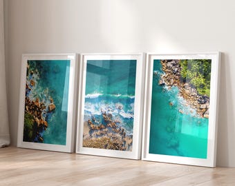 May include: Three framed art prints featuring aerial views of a turquoise ocean with rocky coastlines and white-capped waves. The prints are displayed on a light wood floor, leaning against a white wall.