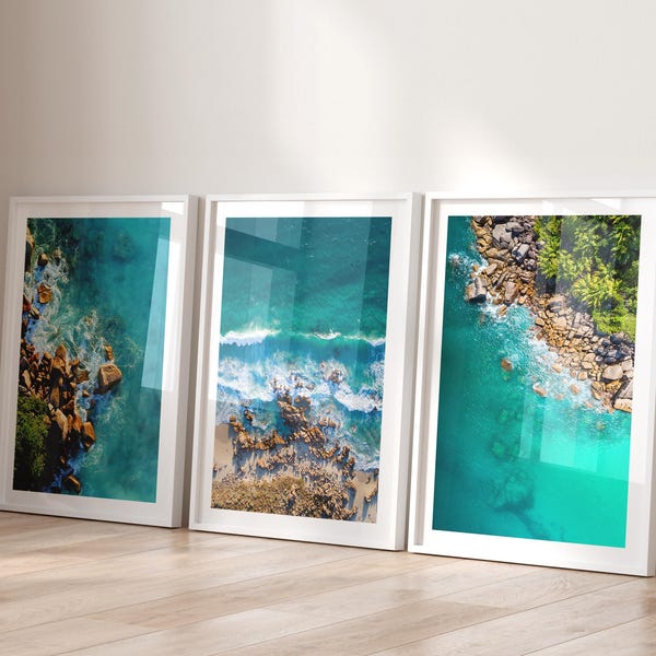 May include: Three framed art prints featuring aerial views of a turquoise ocean with rocky coastlines and white-capped waves. The prints are displayed on a light wood floor, leaning against a white wall.