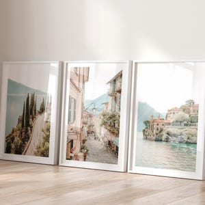May include: Three framed landscape prints. The prints show scenic views of a coastal road, a European street, and a lakeside village. The frames are white, and the images have a soft, muted colour palette.