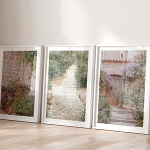 May include: Three framed art prints showcasing European landscapes. The prints depict a stone pathway, a rustic doorway, and a stone wall with greenery. The frames are white, and the overall aesthetic is soft and inviting.