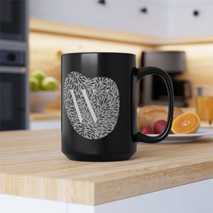 May include: Black ceramic mug with a large handle. The mug features a heart-shaped design filled with a grey floral pattern and a large white letter "W" in the centre. The mug is on a wooden surface.