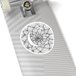 Puede incluir: Una pegatina redonda con la letra "C" blanca sobre un patrón floral en blanco y negro. La pegatina está sobre una tabla de skate con un diseño de rayas grises y blancas.