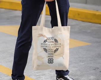 Könnte beinhalten: Beige Canvas-Tragetasche mit dem schwarzen Aufdruck "Way Maker, Miracle Worker, Promise Keeper, Light in the Darkness". Die Tasche hat zwei Griffe und wird von einer Person gehalten.