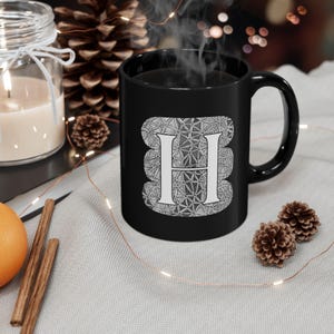 Puede incluir: Una taza de cerámica blanca con asa negra y el texto "But First, Coffee" en negrita negra. La taza se encuentra sobre un fondo borroso, sugiriendo un ambiente acogedor.