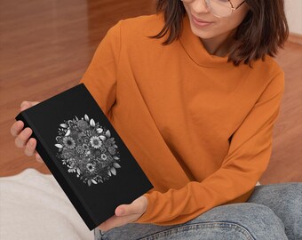 May include: A black book with a detailed floral design in shades of gray and white. The cover features a circular arrangement of various flowers and leaves. The book is held by a person wearing an orange long-sleeve shirt and blue jeans.
