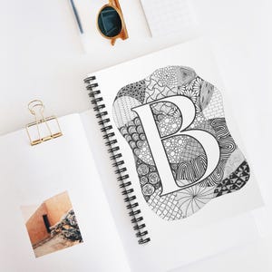 May include: Spiral-bound notebook with a large, white letter "B" in the center, surrounded by a black and white patterned design. The notebook is open, revealing a photo and a gold binder clip. Sunglasses and a pen are also visible.
