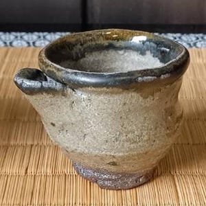 以下が含まれることがあります： ユニークなデザインの、手作りのセラミックカップ3つ。左のカップはひび割れ釉薬のグレー。中央のカップは小さな取っ手付きのタン色。右のカップは赤とグレーのアクセントの白。すべて竹のマットの上にあります。