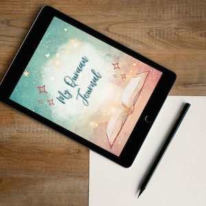 May include: A digital tablet displays a watercolor design with the text "My Quran Journal" and an open book illustration. A black pencil and a blank white paper are on a wooden surface.
