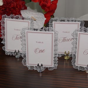 May include: Four rectangular table number cards labeled One, Two, Three, and Four in a dark red script font. Each white card features a thin dark red border and is framed by a delicate white lace trim, held upright by small silver metal stands.