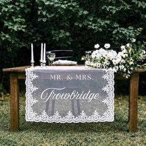 May include: A wooden table draped with a white lace runner featuring the text MR. & MRS. Trowbridge. The table is set with two silver candlesticks, white taper candles, and a large arrangement of white flowers and green foliage on the side.