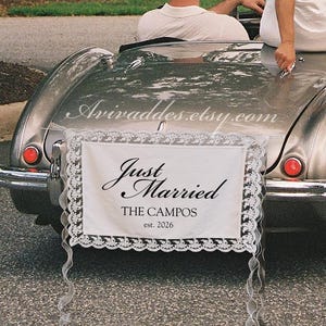 Op de afbeelding: Een zilverkleurige oldtimer cabriolet met een "Just Married" bordje aan de achterkant. Het bordje is wit met zwarte tekst en een kanten rand. Een persoon in een witte jurk zit op de auto.