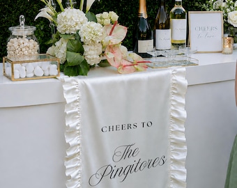 May include: A white satin table runner with ruffled edges, featuring the black printed text "CHEERS TO The Pingitores". The runner is draped over a white bar counter, which is decorated with white floral arrangements, wine bottles, and a sign.