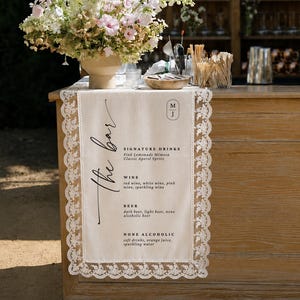 May include: A cream-colored fabric table runner with white lace trim draped over a wooden bar. The runner features the text "the bar," a monogram "M J," and a menu listing signature drinks, wine, beer, and non-alcoholic options in black ink.