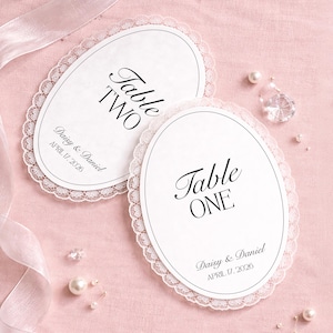 Two white oval table cards with a delicate lace trim, featuring the text "Table ONE" and "Table TWO" in elegant script. The cards also include the names "Daisy & Daniel" and the date "APRIL 17, 2026". The cards are on a pink surface.