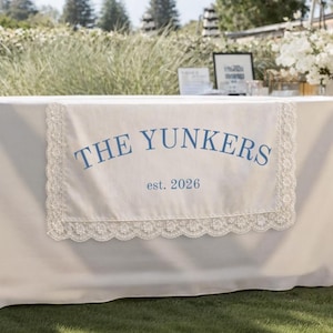 May include: A cream-colored table cloth with a decorative lace trim and the text "THE YUNKERS est. 2026" in blue. The table is set up outdoors on a grassy lawn, with a backdrop of greenery and trees.