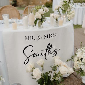 Puede incluir: Una mesa de recepción de bodas con un mantel blanco que muestra el texto "MR. & MRS. Smiths" en escritura negra. La mesa está adornada con rosas blancas, velas y arreglos florales, creando un ambiente elegante y romántico.