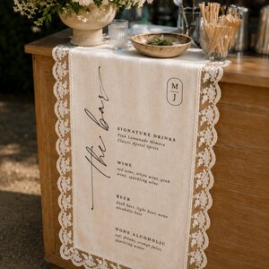 May include: A beige linen table runner with white lace trim featuring black printed text that reads the bar, signature drinks, wine, beer, and none alcoholic, along with a monogram logo featuring the letters M and J in a vertical oval frame.