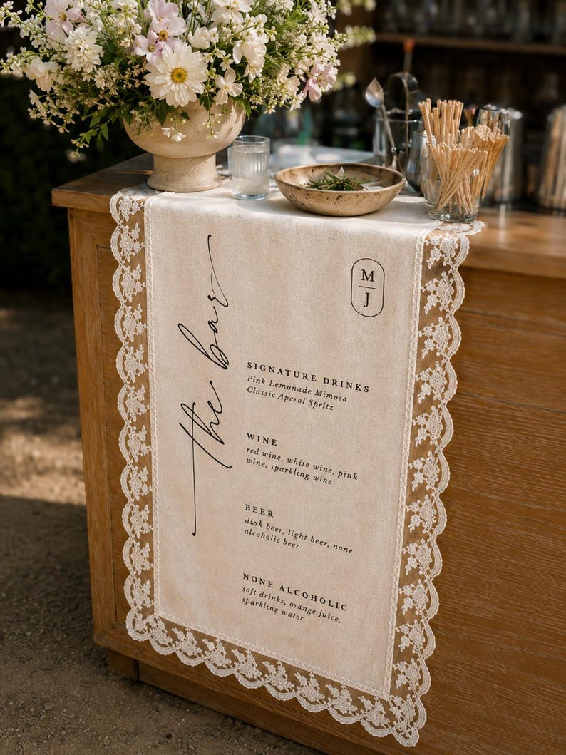 May include: A beige linen table runner with white lace trim featuring black printed text that reads the bar, signature drinks, wine, beer, and none alcoholic, along with a monogram logo featuring the letters M and J in a vertical oval frame.
