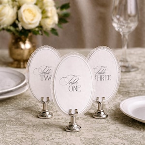 Three elegant table number holders with oval cards, each displaying a table number in script font. The cards have a delicate lace-like border and are held by silver-toned metal stands. A vase of white roses and a wine glass are in the background.