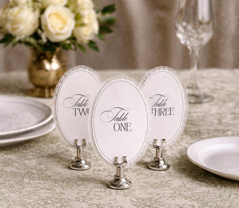 Three elegant table number holders with oval cards, each displaying a table number in script font. The cards have a delicate lace-like border and are held by silver-toned metal stands. A vase of white roses and a wine glass are in the background.