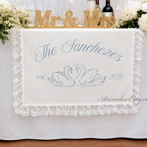 May include: A rectangular off-white fabric table runner featuring a blue embroidered design of two swans facing each other. The text reads The Sanchezes, est. 2021, and Avivades.Etsy.com. The runner is bordered by a wide, white scalloped lace trim.
