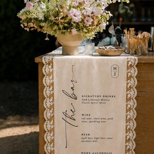May include: A beige fabric table runner with white lace trim featuring the printed text The bar, signature drinks, wine, beer, and none alcoholic options. The runner is displayed on a wooden surface with a large floral arrangement in a beige vase.