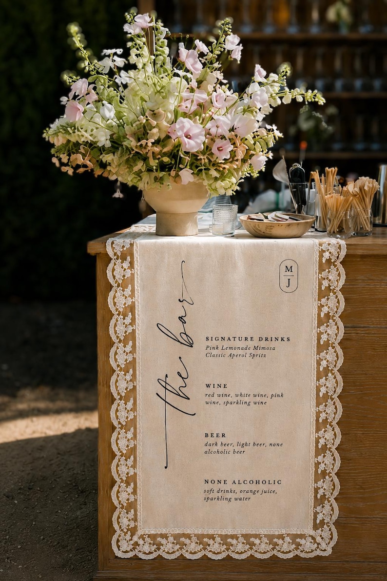 May include: A beige fabric table runner with white lace trim featuring the printed text The bar, signature drinks, wine, beer, and none alcoholic options. The runner is displayed on a wooden surface with a large floral arrangement in a beige vase.