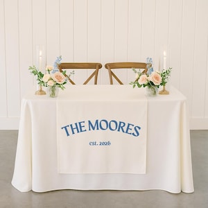 Puede incluir: Una mesa color crema con un mantel blanco y un camino de mesa impreso con "THE MOORES est. 2026" en azul. Dos sillas de madera están detrás de la mesa. Arreglos florales y velas decoran la mesa.
