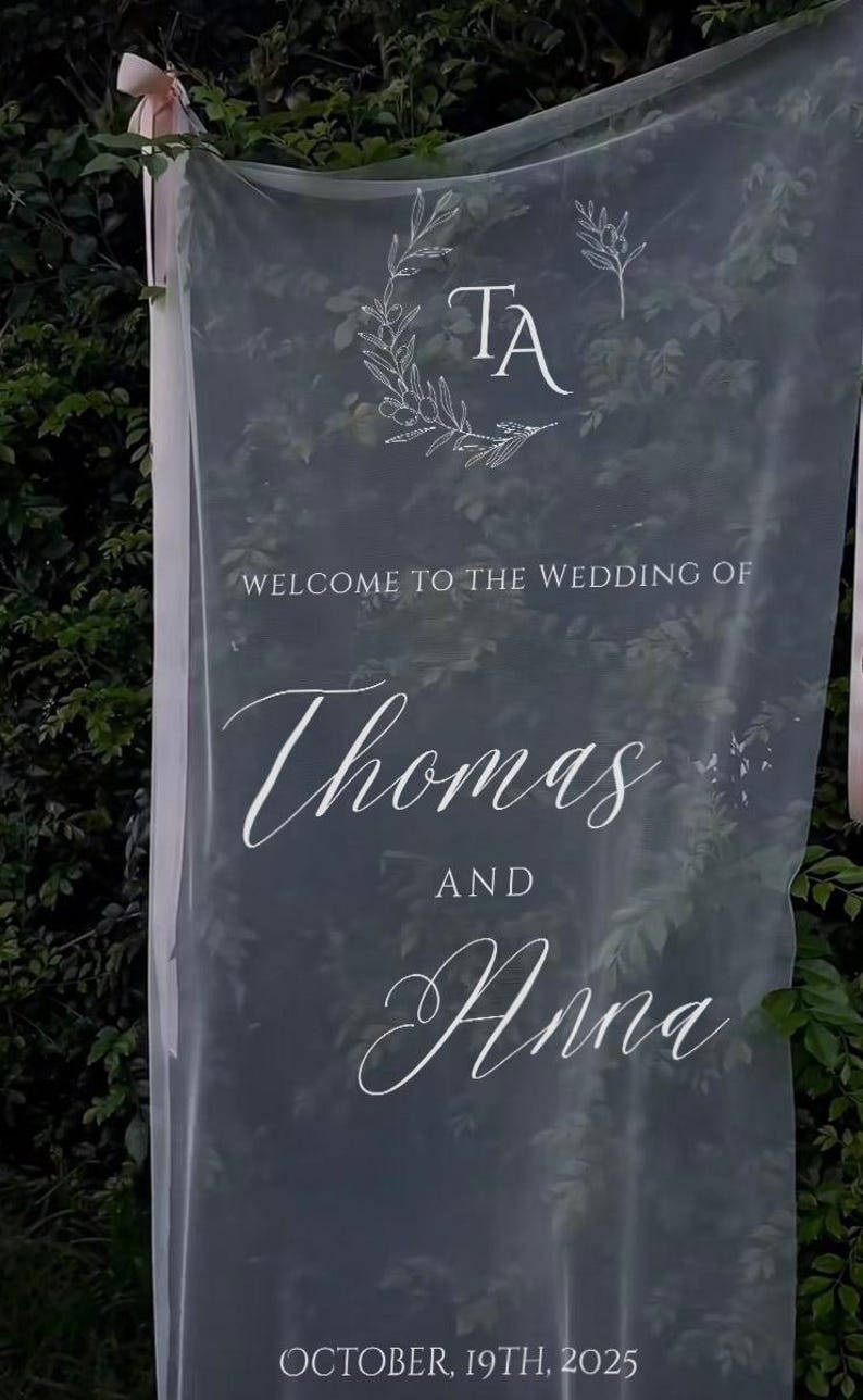 Puede incluir: Cartel de boda de tela transparente con texto blanco. El cartel dice "BIENVENIDOS A LA BODA DE Thomas Y Anna" y "19 DE OCTUBRE DE 2025". La parte superior del cartel tiene las iniciales "TA" dentro de una corona floral.
