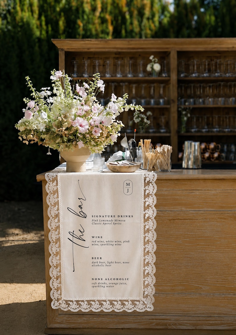 May include: A cream-colored fabric table runner with white lace trim draped over a wooden bar. The runner features the text "the bar," a monogram "M J," and a menu listing signature drinks, wine, beer, and non-alcoholic options in black ink.