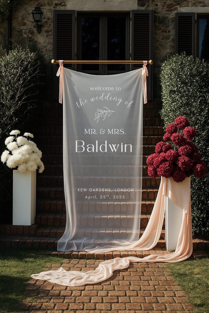Puede incluir: Decoraci&oacute;n de ceremonia de boda con una pancarta blanca transparente con el texto "welcome to the wedding of MR. & MRS. Baldwin". La pancarta est&aacute; sobre una escalera, flanqueada por arreglos florales y tela.