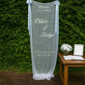 Puede incluir: Un letrero de bienvenida de boda blanco transparente con cintas azul claro. El letrero dice "Bienvenida a la boda de Oliver & Evelyn" e incluye la fecha y el lugar: Cedar Lakes Estate, Nueva York, 17 de marzo de 2025.