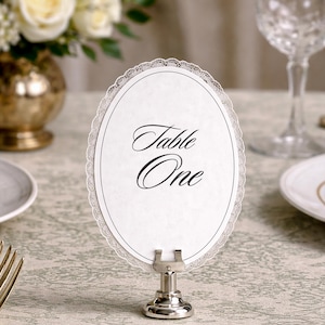 An oval table number card with the words "Table One" in elegant script. The white card has a delicate lace trim and is held by a silver metal stand. The setting includes a floral arrangement and glassware.
