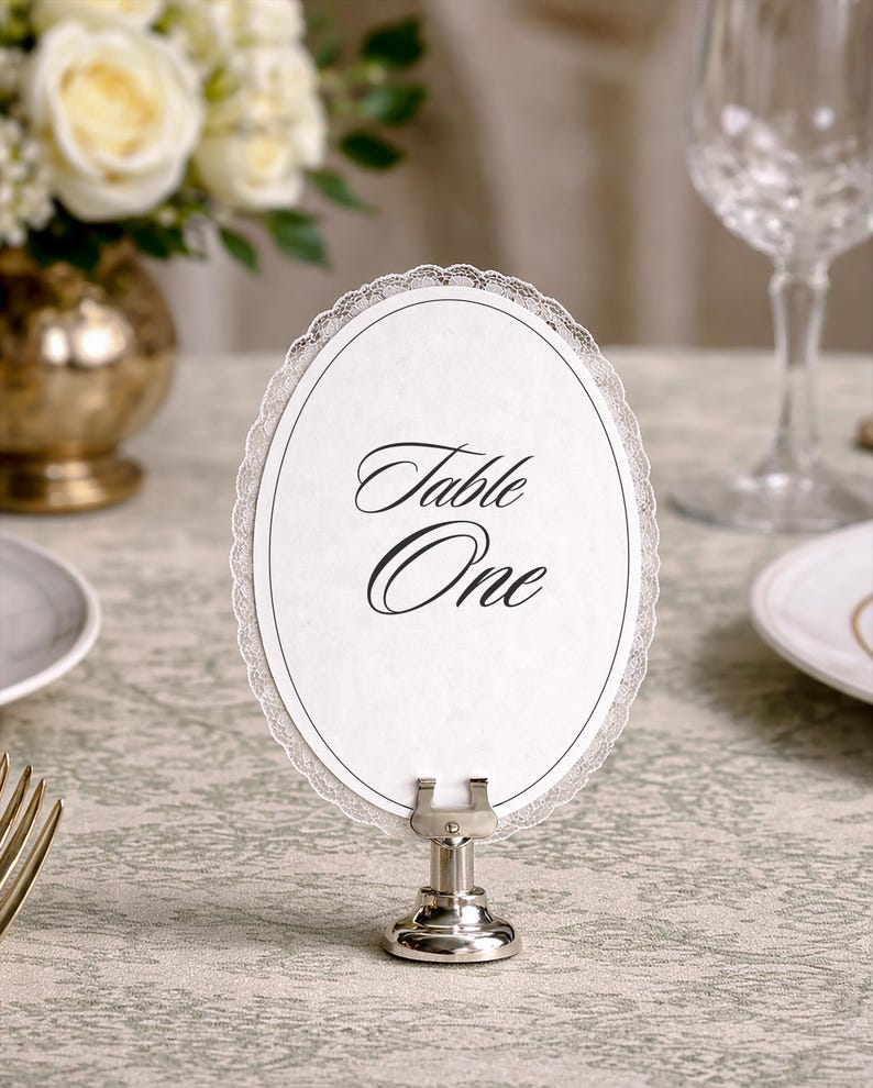 An oval table number card with the words "Table One" in elegant script. The white card has a delicate lace trim and is held by a silver metal stand. The setting includes a floral arrangement and glassware.