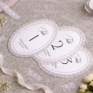 Oval-shaped table number cards with a delicate lace trim. Each card features a bold number and the word "Table" in elegant script. The cards are arranged on a textured surface, with floral accents and a lit candle.