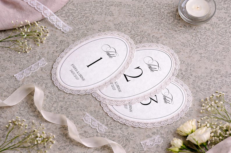 Oval-shaped table number cards with a delicate lace trim. Each card features a bold number and the word "Table" in elegant script. The cards are arranged on a textured surface, with floral accents and a lit candle.