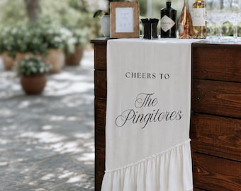 May include: A cream-colored fabric table runner featuring a ruffled bottom edge and black printed text that reads "CHEERS TO The Pingitores". The runner is draped over a dark brown wooden bar counter, with bottles and a framed sign visible on top.
