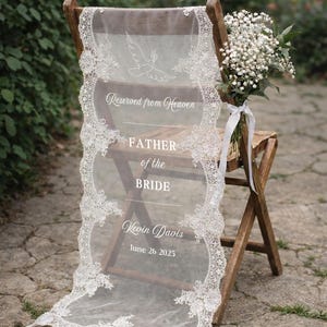 May include: A sheer white fabric memorial sign draped over a wooden folding chair, featuring intricate lace borders, a dove illustration, and white text reading Reserved from Heaven, FATHER of the BRIDE, Kevin Davis, and the date June 26 2025.