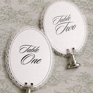 Two oval-shaped table place cards with a delicate lace trim. Each card has the words "Table One" and "Table Two" in elegant script. The cards are held by silver metal stands, perfect for weddings or special events.