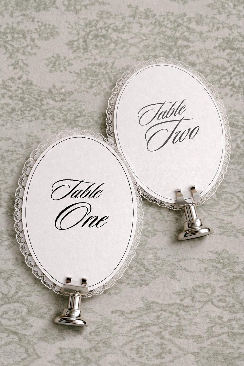 Two oval-shaped table place cards with a delicate lace trim. Each card has the words "Table One" and "Table Two" in elegant script. The cards are held by silver metal stands, perfect for weddings or special events.