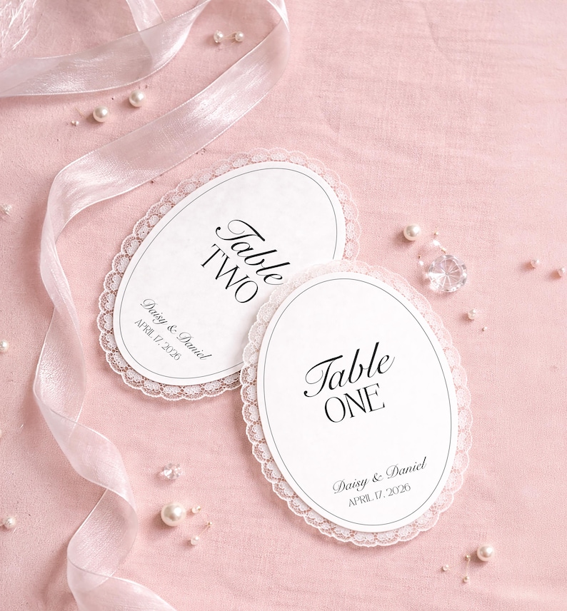 Two white oval table cards with a delicate lace trim, featuring the text "Table ONE" and "Table TWO" in elegant script. The cards also include the names "Daisy & Daniel" and the date "APRIL 17, 2026". The cards are on a pink surface.
