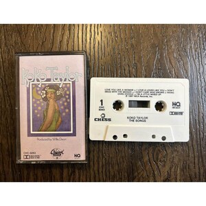 May include: A vintage cassette tape of Koko Taylor's album, "The Songs." The cassette case is light purple with an illustration of a woman. The white cassette tape has song titles and the Chess Records logo.