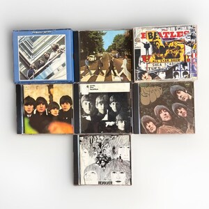 May include: A collection of eight compact discs featuring album art from The Beatles. The album covers display various images, including the band members, street scenes, and abstract designs. The CDs are arranged on a white surface.