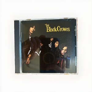 May include: A compact disc in a clear plastic jewel case featuring a dark cover with a group of people in shadows. The text The Black Crowes is printed in yellow in the upper right corner. The case has a black spine and a transparent back.